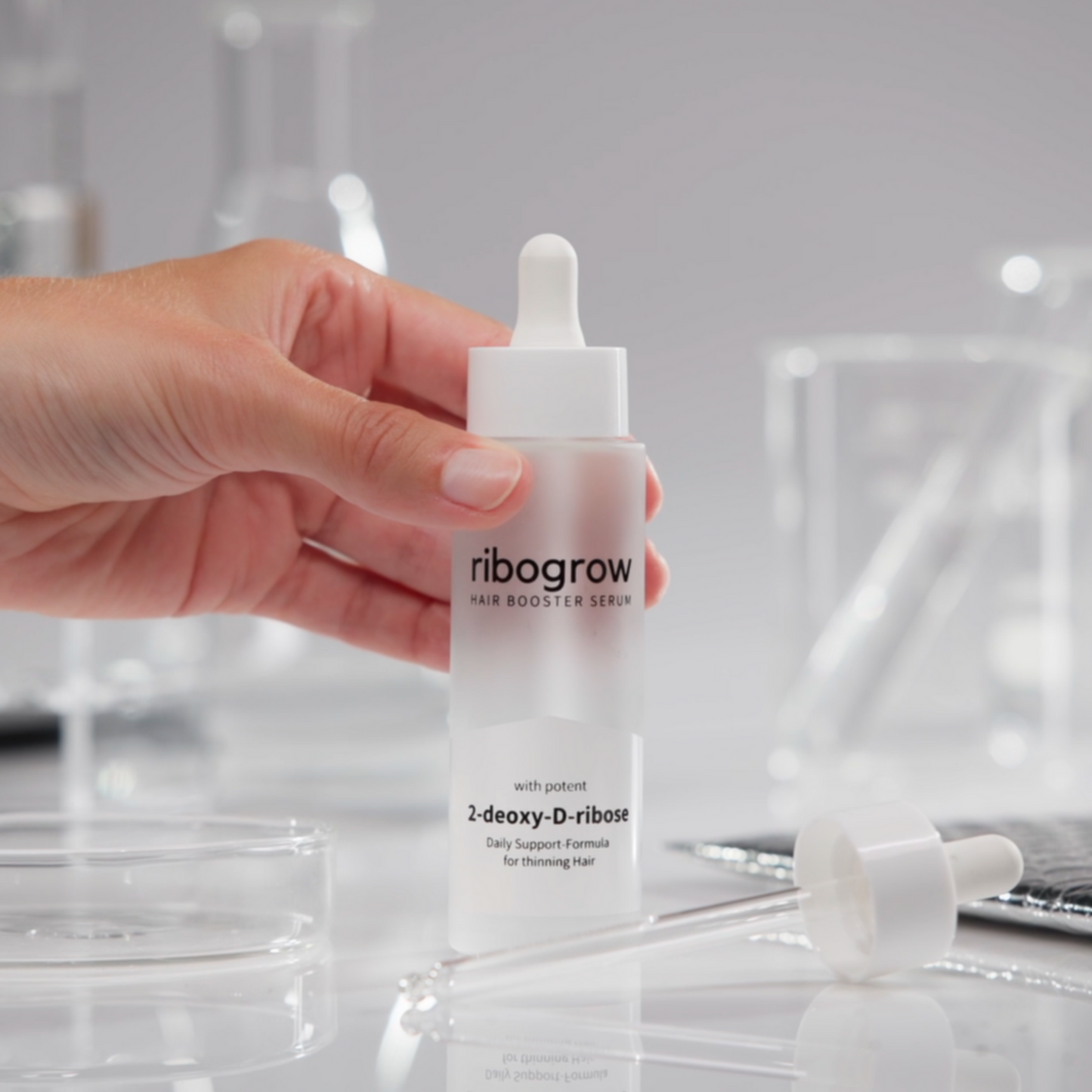 Nahaufnahme einer Hand, die das ribogrow Hair Booster Serum mit Pipette hält. Hochwertiges Haarserum mit 2-Desoxy-D-Ribose zur täglichen Unterstützung bei dünner werdendem Haar. Elegantes, minimalistisches Produktfoto mit wissenschaftlichem Hintergrund.