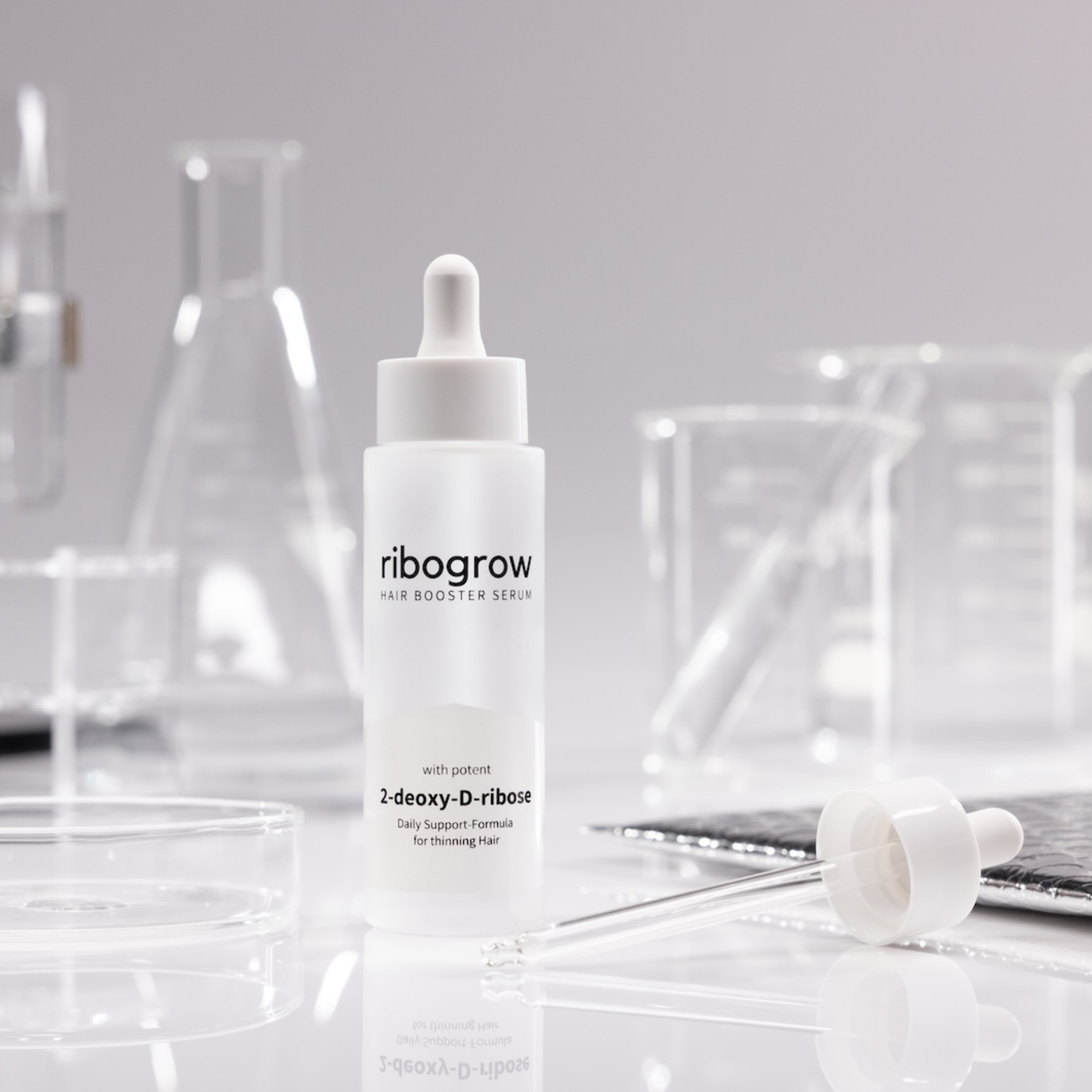 Flasche ribogrow Hair Booster Serum aus mattem Glas mit Pipettenverschluss, stehend auf weißer Oberfläche, umgeben von Labor-Glaswaren wie Bechern, Petrischalen und Reagenzgläsern in einem wissenschaftlich anmutenden Setting.