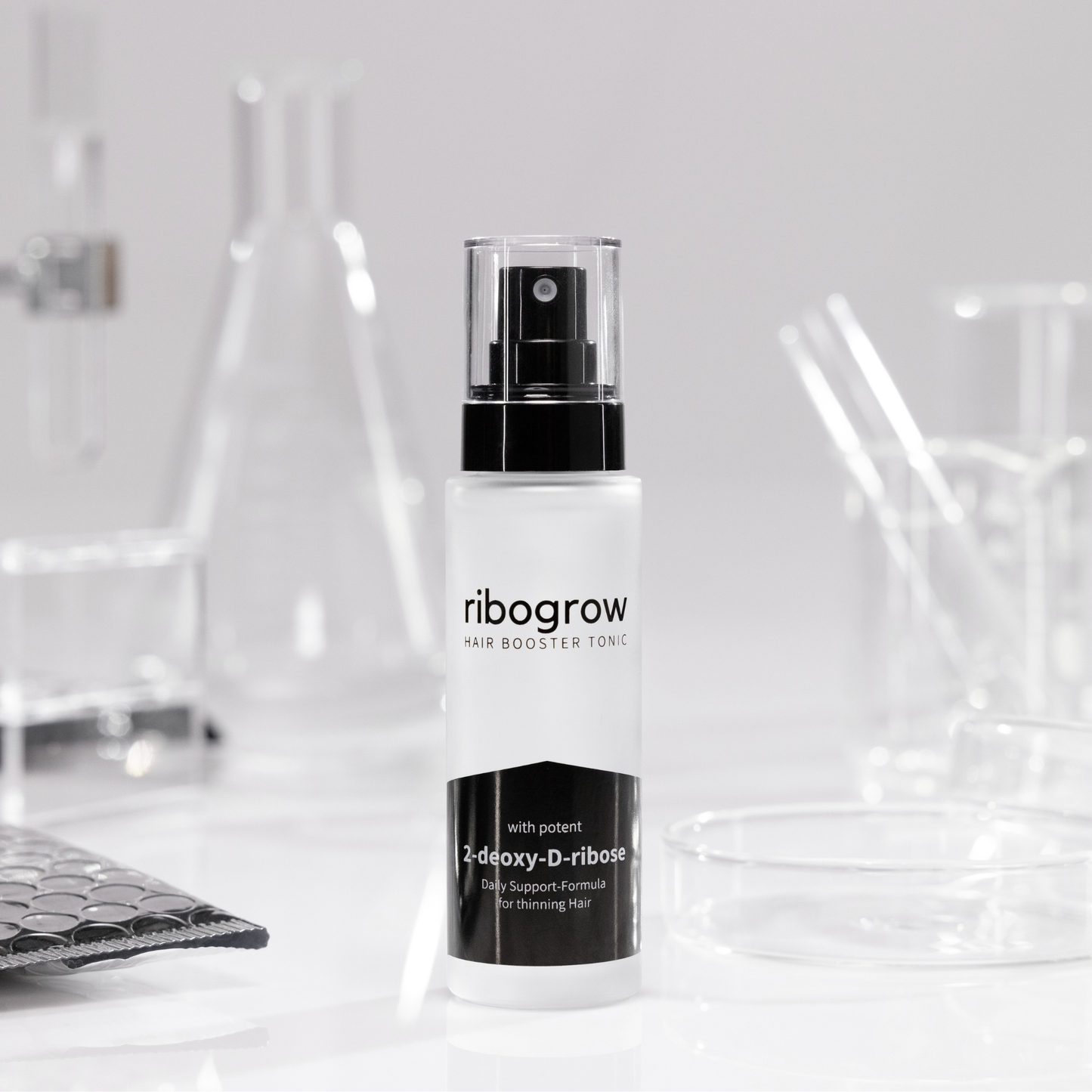 Flasche ribogrow Hair Booster Tonic aus mattem Glas mit schwarzem Sprühkopf, stehend auf weißer Oberfläche, umgeben von Labor-Glaswaren wie Bechern, Petrischalen und Reagenzgläsern in einem wissenschaftlich inspirierten Umfeld.