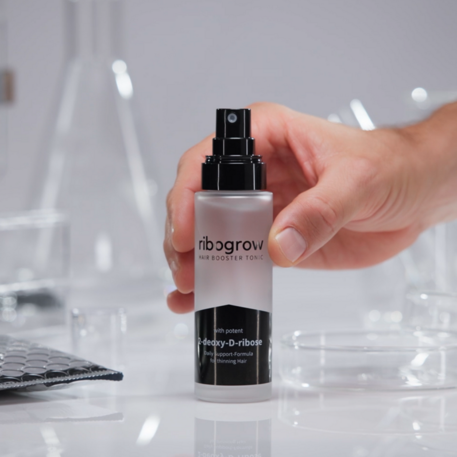 Nahaufnahme einer Hand, die das ribogrow Hair Booster Tonic mit Sprühkopf hält. Hochwertiges Haar-Tonic mit 2-Desoxy-D-Ribose zur täglichen Unterstützung bei Haarausfall und dünner werdendem Haar. Modernes, minimalistisches Produktfoto mit Labor-Setting.