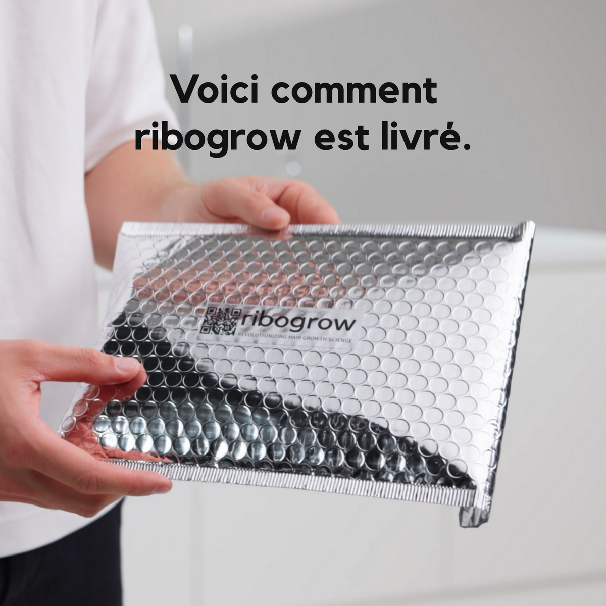 Une personne tient une enveloppe d’expédition argentée à bulles, ornée du logo ribogrow et d’un QR code. L’image présente l’emballage de livraison du ribogrow Hair Booster Tonic, conçu pour protéger le produit pendant le transport.