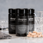 Drei schwarze Dosen ribogrow® Hair Booster Capsules mit glänzenden Schraubdeckeln und grau-schwarzen Etiketten, angeordnet auf einer hellen, spiegelnden Oberfläche. Im Vordergrund steht eine Glasschale mit hellbeigen, orange gesprenkelten Kapseln. Transparente Laborgefäße und Laborutensilien im Hintergrund unterstreichen die wissenschaftliche Produktpräsentation.