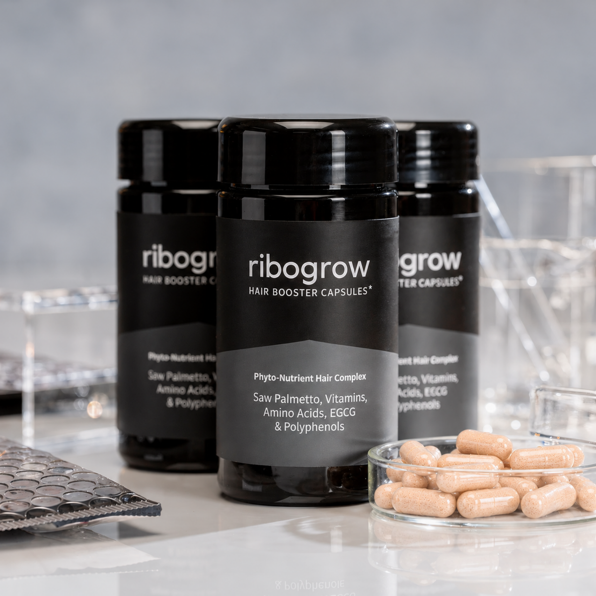 Drei schwarze Dosen ribogrow® Hair Booster Capsules mit glänzenden Schraubdeckeln und grau-schwarzen Etiketten, angeordnet auf einer hellen, spiegelnden Oberfläche. Im Vordergrund steht eine Glasschale mit hellbeigen, orange gesprenkelten Kapseln. Transparente Laborgefäße und Laborutensilien im Hintergrund unterstreichen die wissenschaftliche Produktpräsentation.