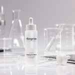 Flasche ribogrow Hair Booster Serum aus mattem Glas mit Pipettenverschluss, stehend auf weißer Oberfläche, umgeben von Labor-Glaswaren wie Bechern, Petrischalen und Reagenzgläsern in einem wissenschaftlich anmutenden Setting.