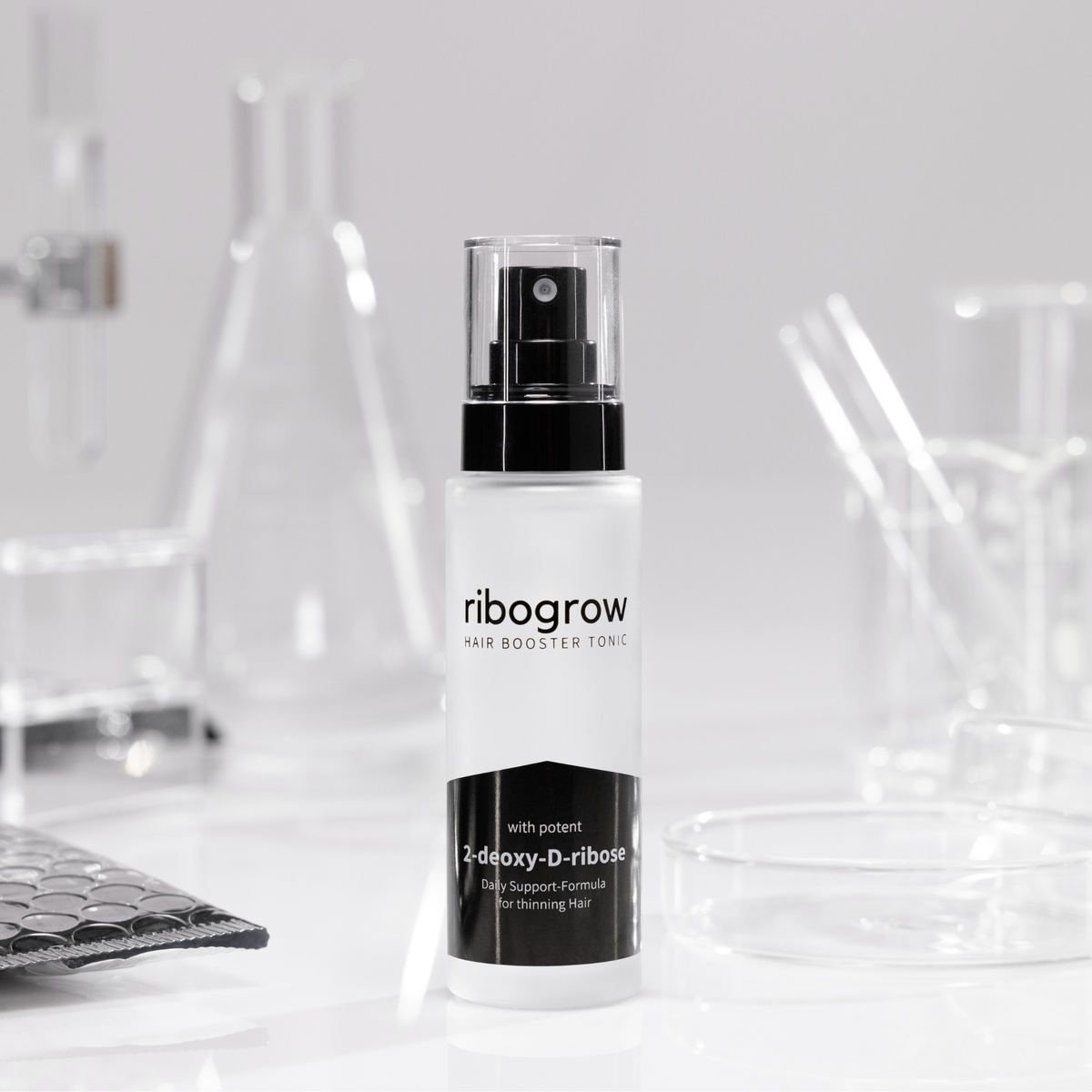 Flasche ribogrow® Hair Booster Tonic aus mattem Glas mit schwarzem Sprühkopf, stehend auf weißer Oberfläche, umgeben von Labor-Glaswaren wie Bechern, Petrischalen und Reagenzgläsern in einem wissenschaftlich inspirierten Umfeld.