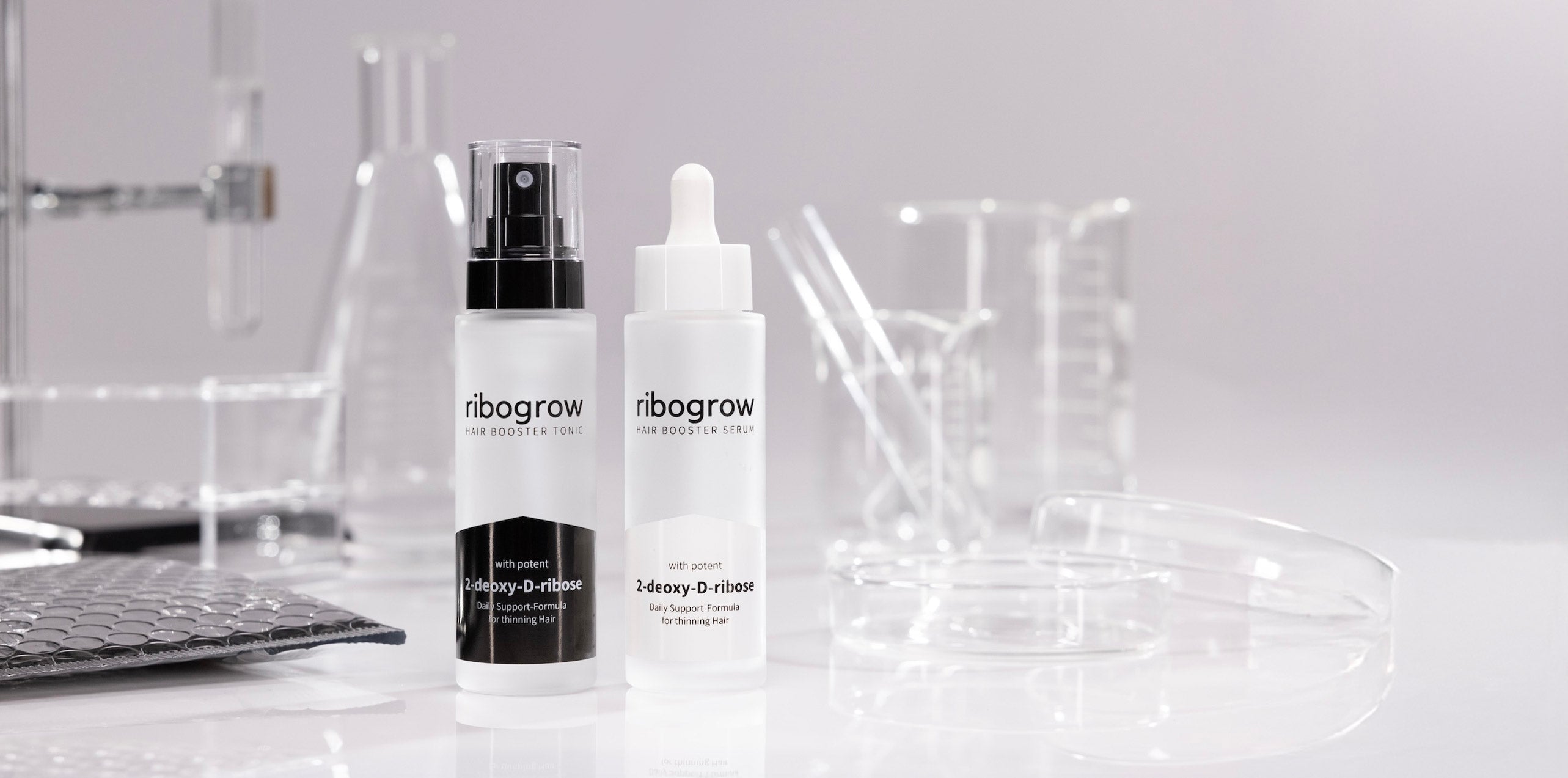 Two frosted bottles of ribogrow hair booster products, one tonic with a spray pump and one serum with a dropper, placed on a white surface surrounded by transparent laboratory glassware and petri dishes in a clean, scientific setting.
