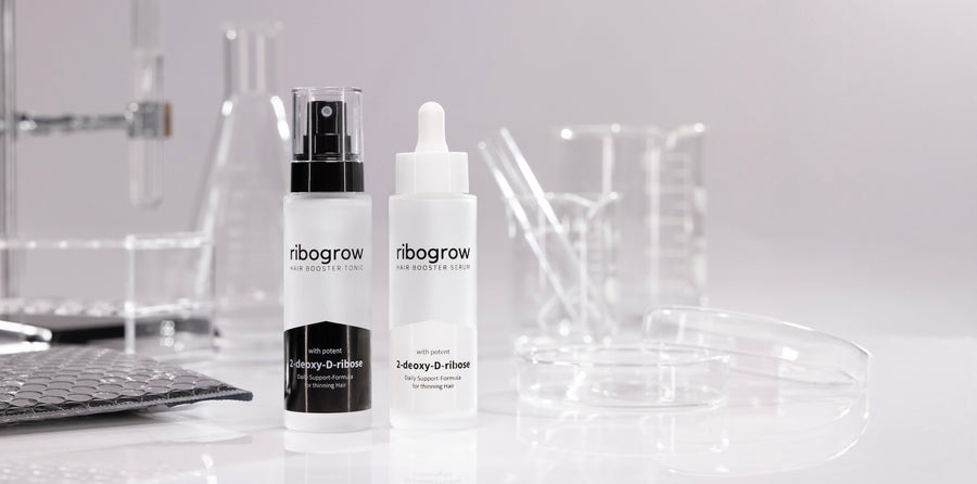 Two frosted bottles of ribogrow hair booster products, one tonic with a spray pump and one serum with a dropper, placed on a white surface surrounded by transparent laboratory glassware and petri dishes in a clean, scientific setting.
